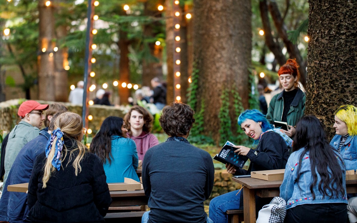 Image of a group of young people sitting on picnic tables looking to one person who's smiling, reading the Perth Festival brochure.