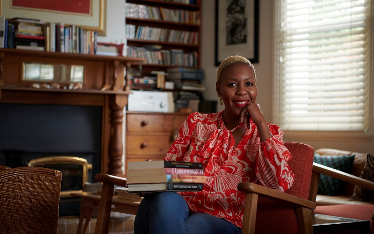 Image of a woman (Sisonke Msimang) in a red and white floral print shirt, seated with a stack of books in front of her.