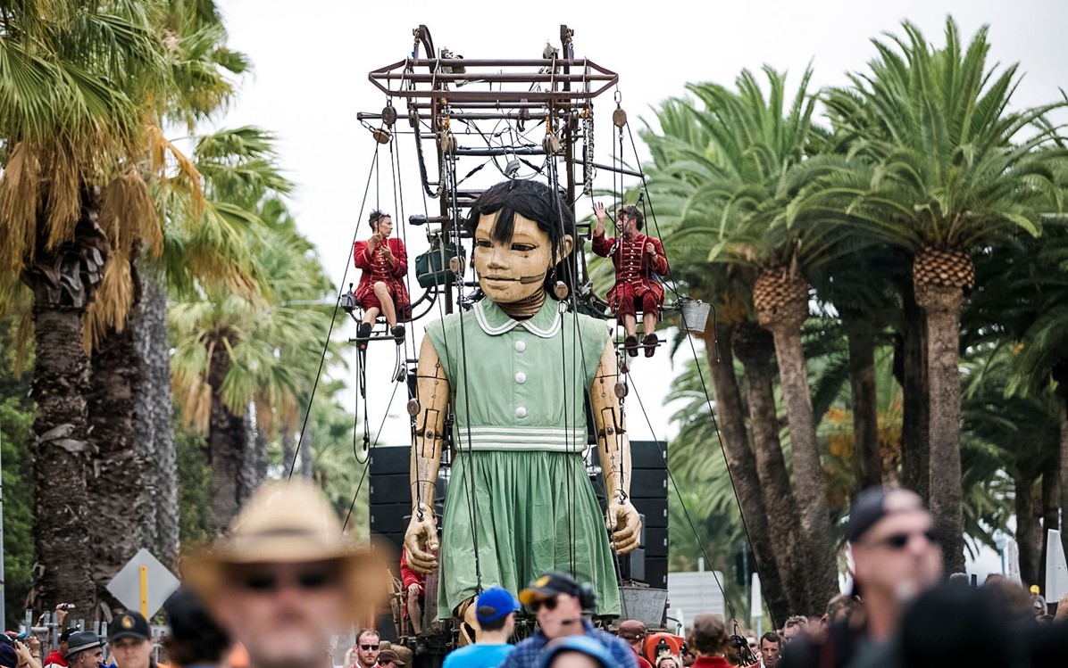 Image of a the girl puppet from the 2015 Perth Festival event 'The Giants', amongst palm trees and crowds, puppeteers on either shoulder.