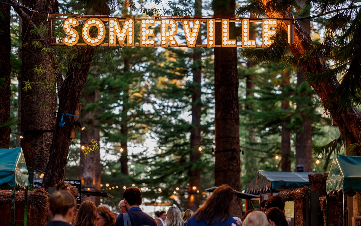 Lit up Somerville sign amongst trees at entrance to outdoor cinema. People walking in. 
