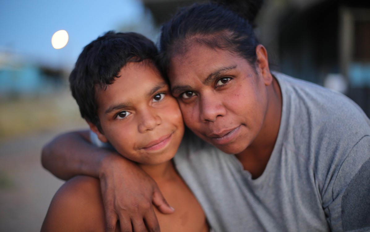 An Aboriginal mother and eight year old son smile and hug each other looking at the camera. 