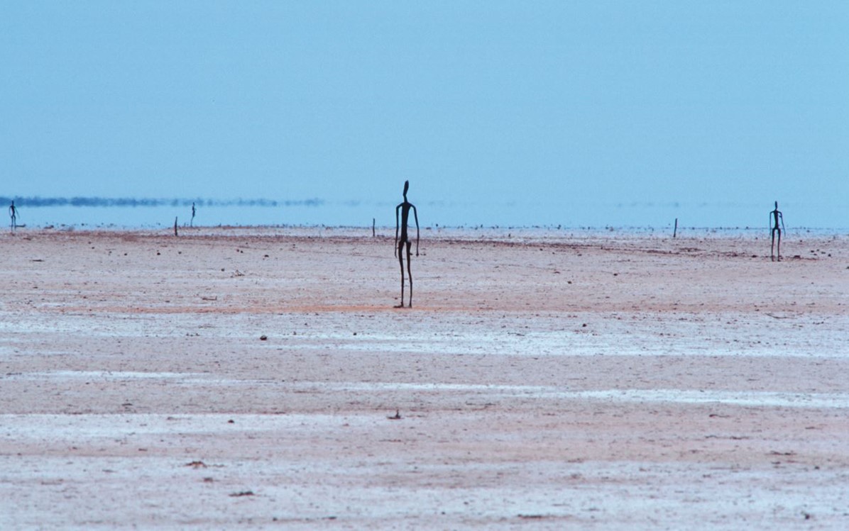 An image of a dark humanlike sculpture on a sand field with a few other sculptures in the background