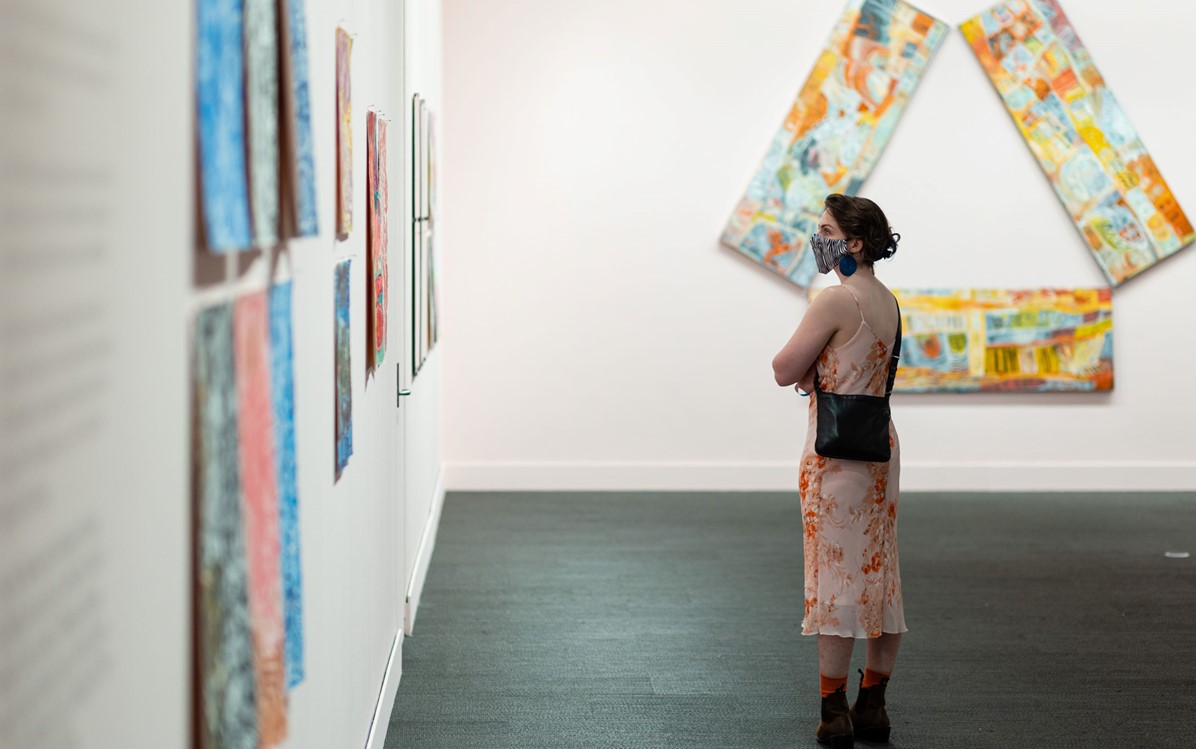 A woman in a patterned slip dress stands with her arms crossed looking at artworks on a wall in a gallery.