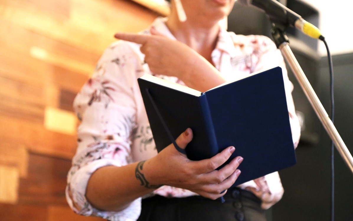 A woman is pictured at a microphone holding a navy blue notebook in one hand, and pointing with the other.