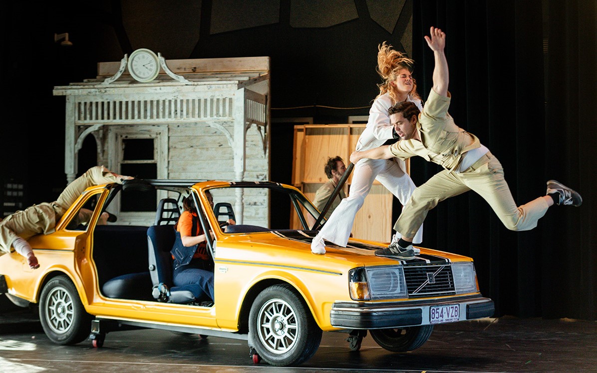 Two performers in action poses standing on the bonnet of an old yellow car with no doors. Person is sat in the drivers seat and scene set of white house is in the background. 