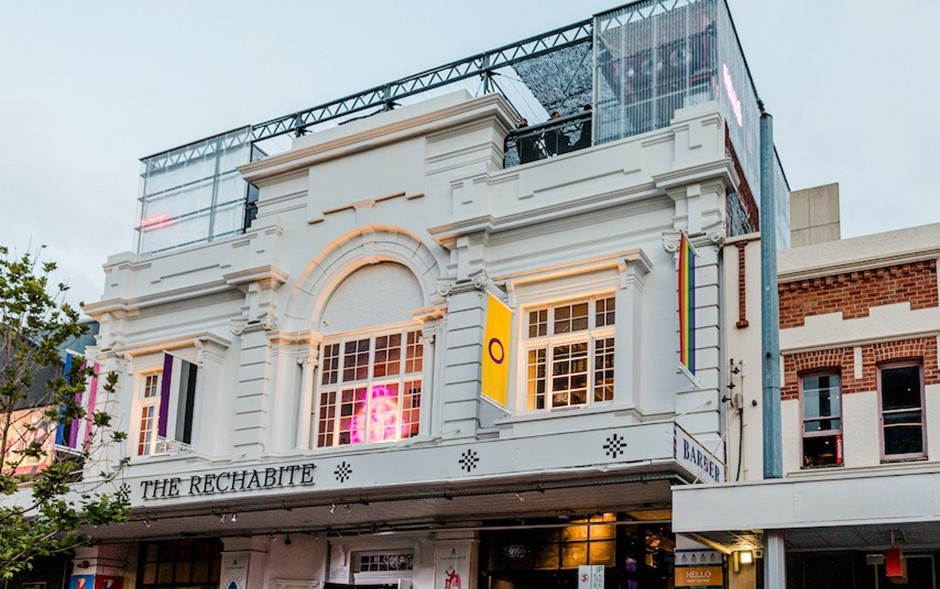 An image of the exterior of The Rechabite. An historic building of a white colour with neon lights coming through the windows
