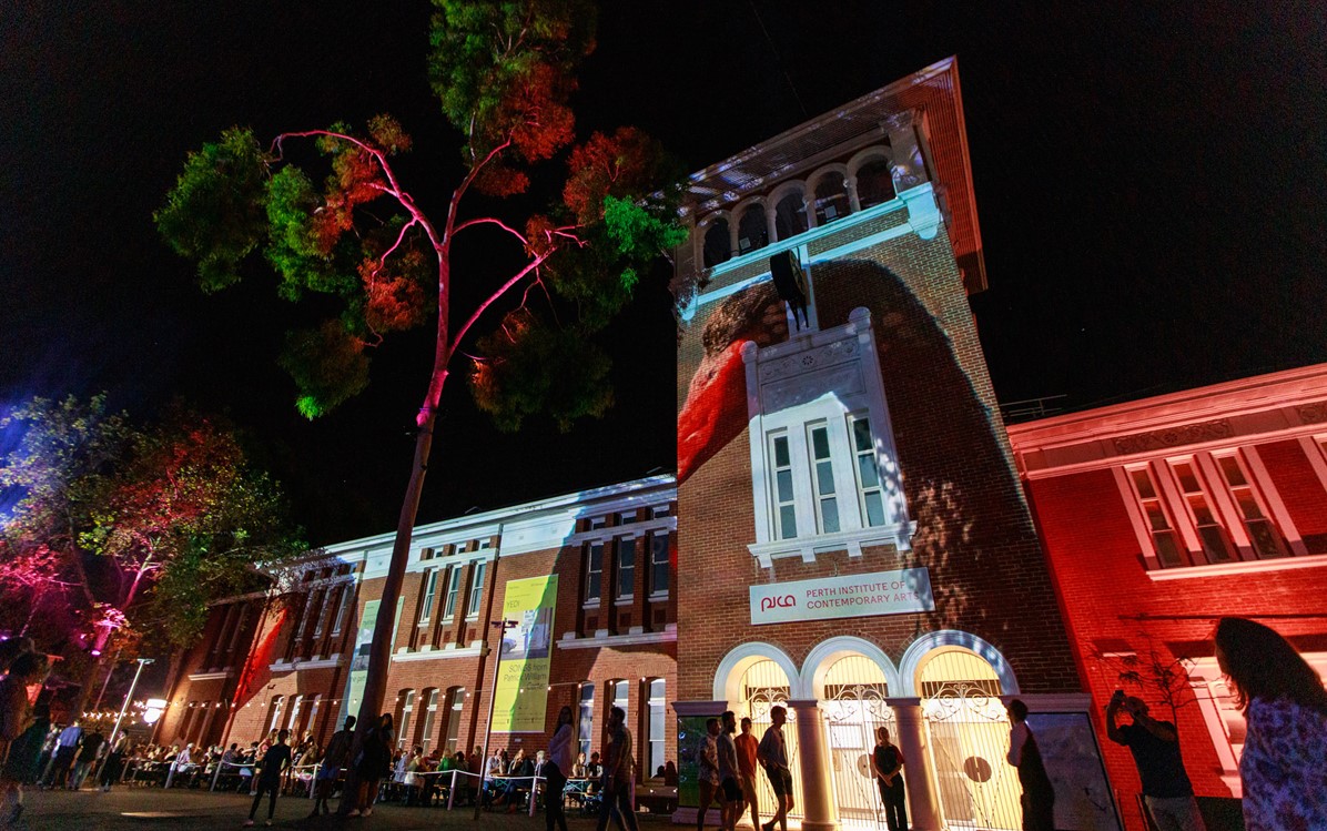 The external of PICA Art Gallery at night with colourful projections on the external walls and trees in front of the building
