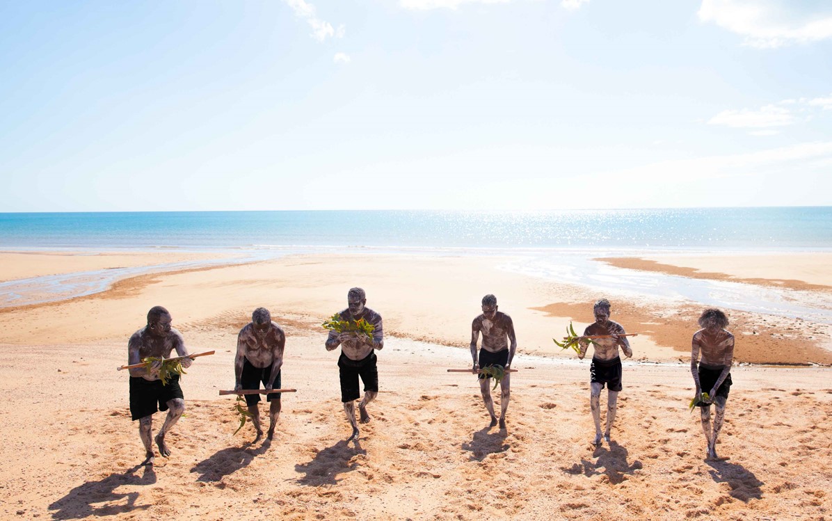 Image of 6 indigenous men standing on the shore and each holding a pole with some leaves from ''Bunggul''