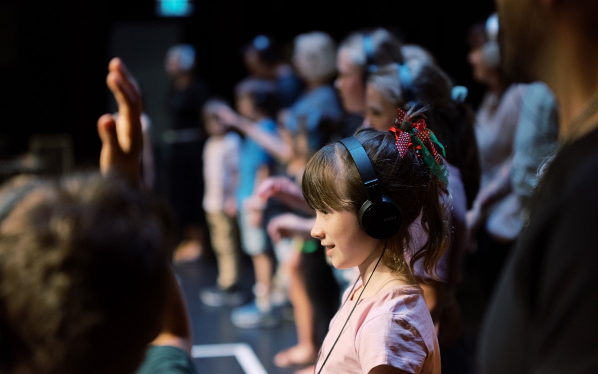 A young girl in a pink top has headphones on and is looking at a stage. More children and adults are out of focus in the background standing up with headphones on.