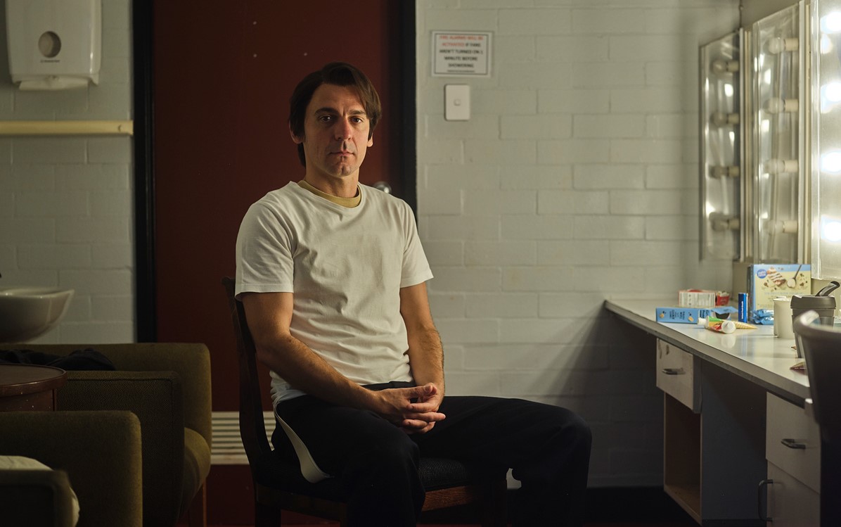 A full body portrait of a man seated in a dressing room. They have a straight expression and is looking straight at the camera.  Credit Duncan Wright