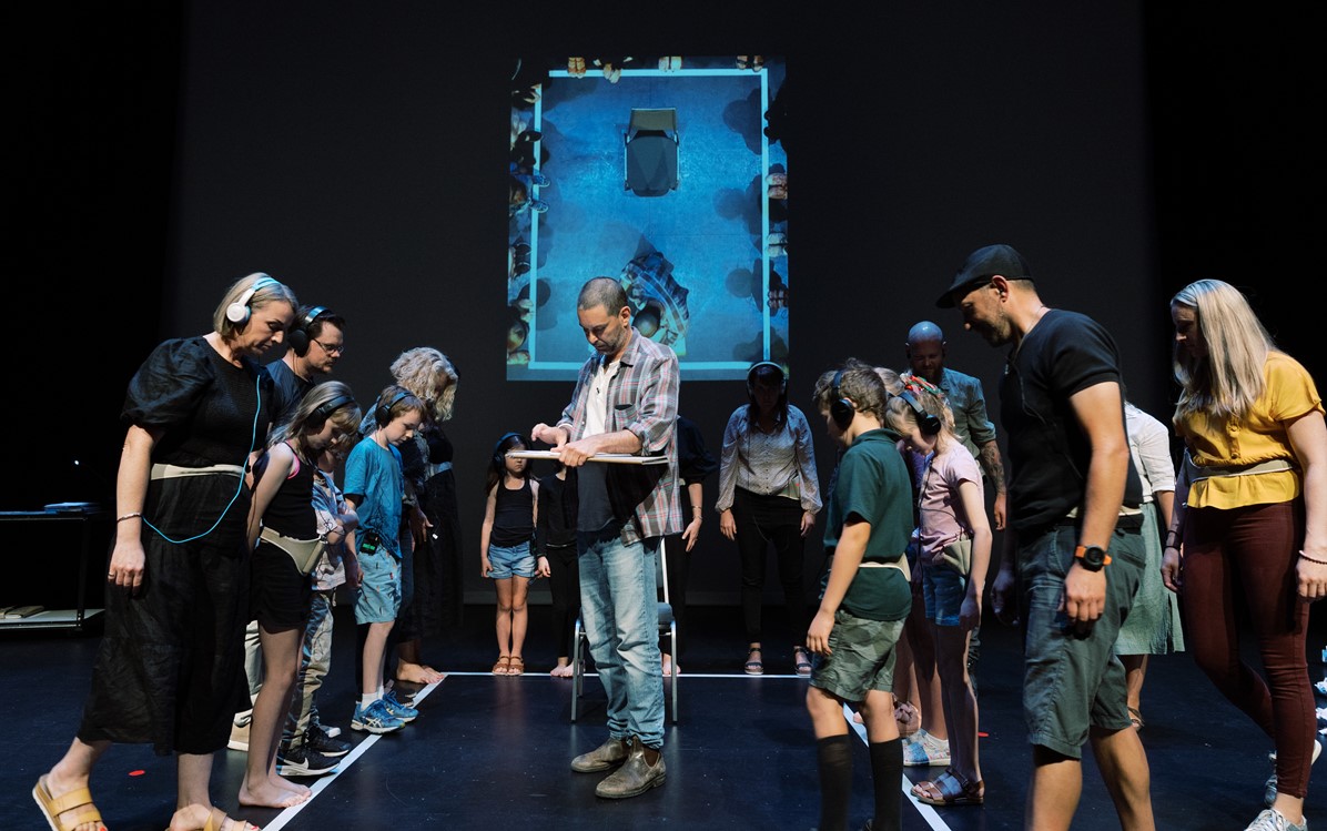 Ben Mortley stars in Kim Crotty's play the Smallest Stage. He's in the centre of a stage, surrounded by adults and children with headphones on. He's drawing on a board, and a bird's eye view of the stage is projected on the back wall.