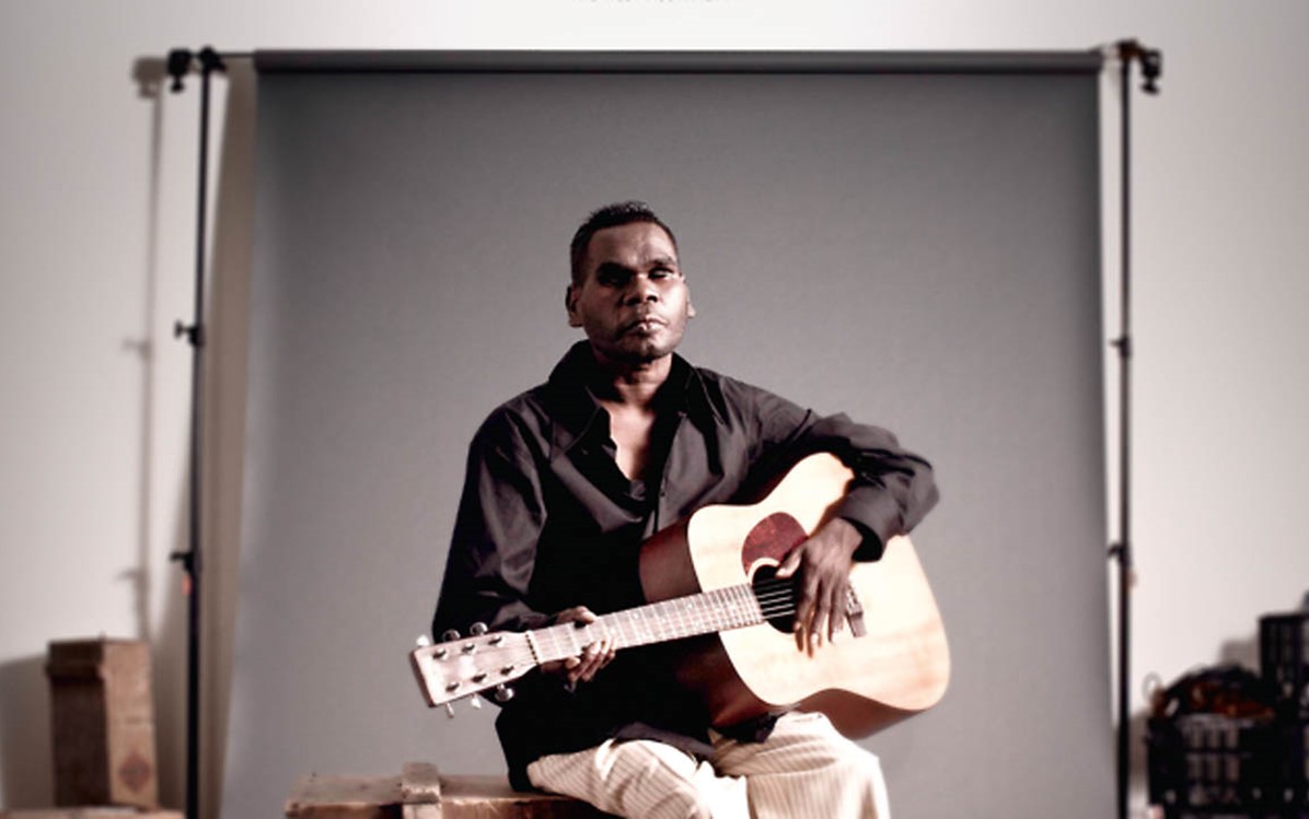 A man (Gurrumul) in a black shirt is pictured holding an acoustic guitar.