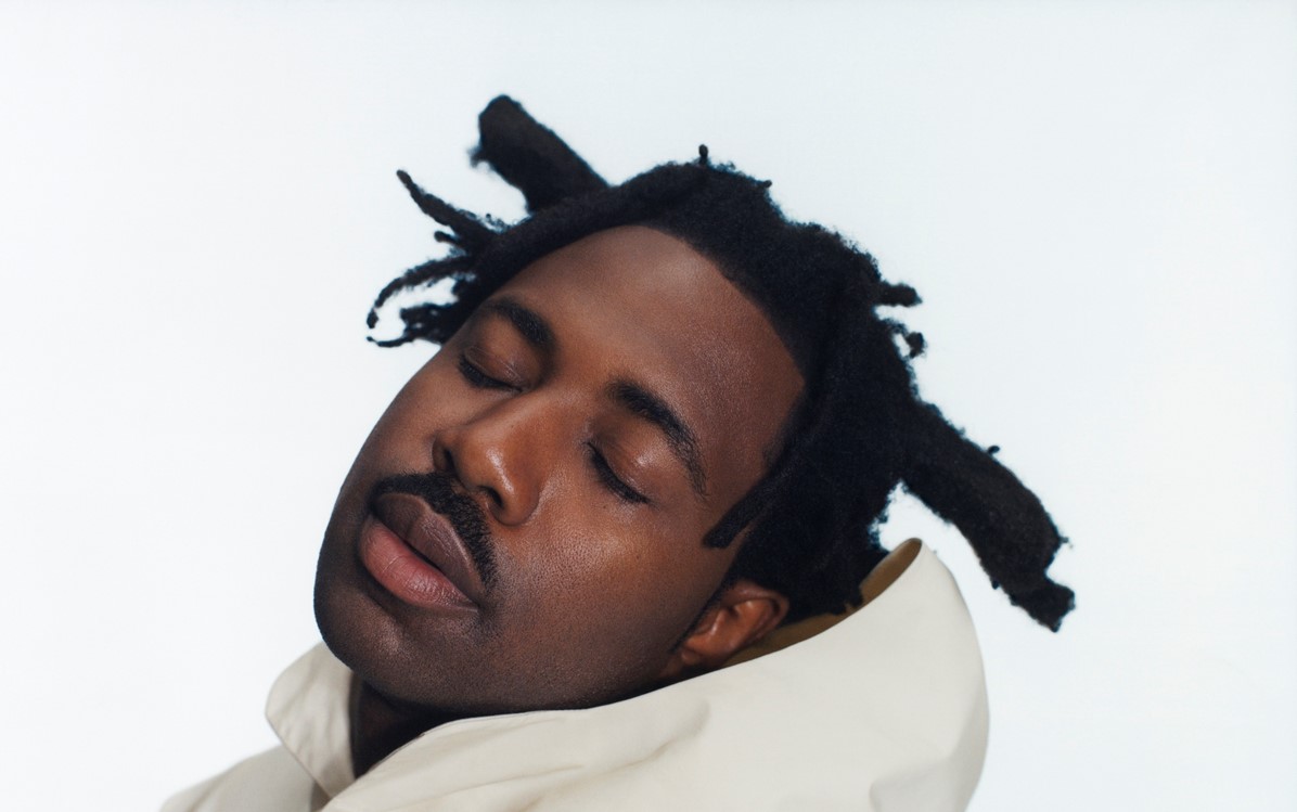 Sampha leaning to the right with their eyes closed in a white hooded jumper