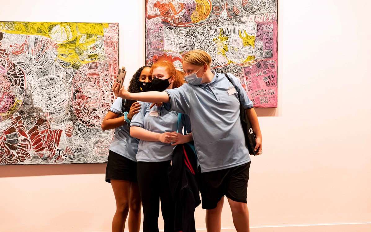 A group of 3 school children take a selfie in front of paintings by Sonia Kurarra, at Lawrence Wilson Art Gallery.