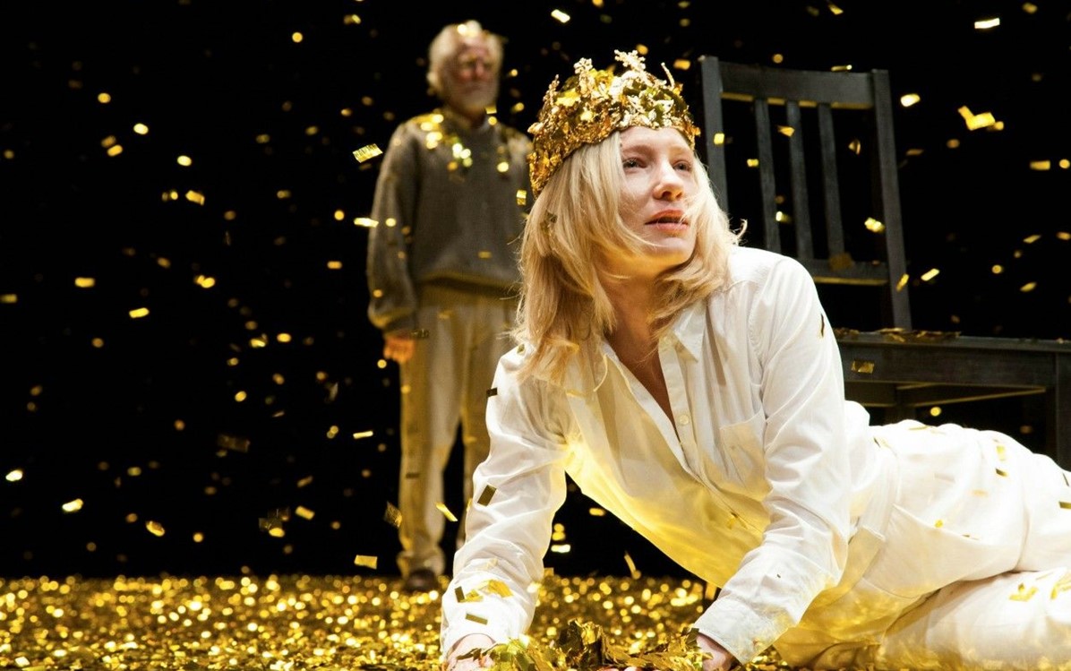 An image of a performer lying on their side propped up on one arm in a white outfit, a gold crown and lying on a stage covered in gold confetti
