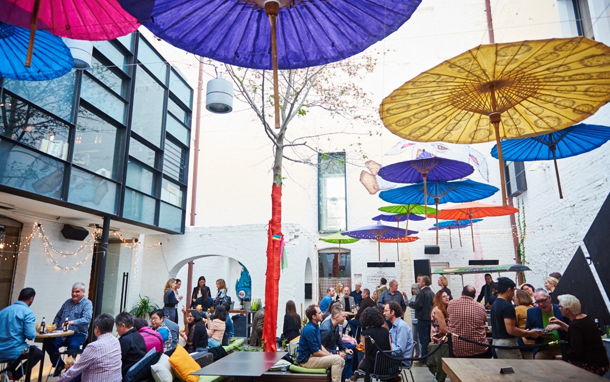 Many people in a courtyard with varied seating and brightly coloured paper umbrellas hanging above.