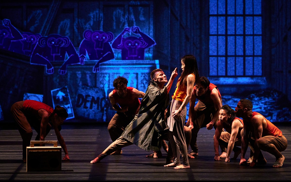 Performers on stage with blue background lighting. Main performer lunging and pointing at another performer who's standing looking at main performer. 5 other performers crouched around. l