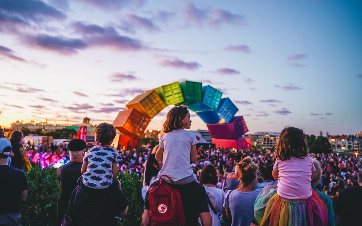 An image from Highway to Hell, three children on adults shoulders are in the foreground with a huge crowd congregates as the sun sets over the rainbow container sculpture in Fremantle