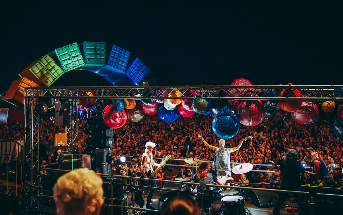 Image of band members on stage and audience members at highway to hell with rainbow shipping container arch in the background
