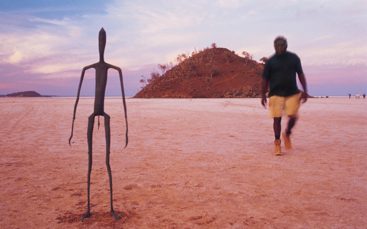 Image of Australian outback desert, a metal sculpture of a man and a blurred man walking towards the camera.