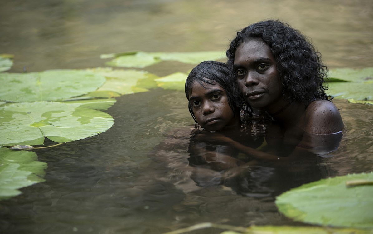 A mother and child are pictured partially submerged in water surrounded by large floating leaves. From the film "High Ground".