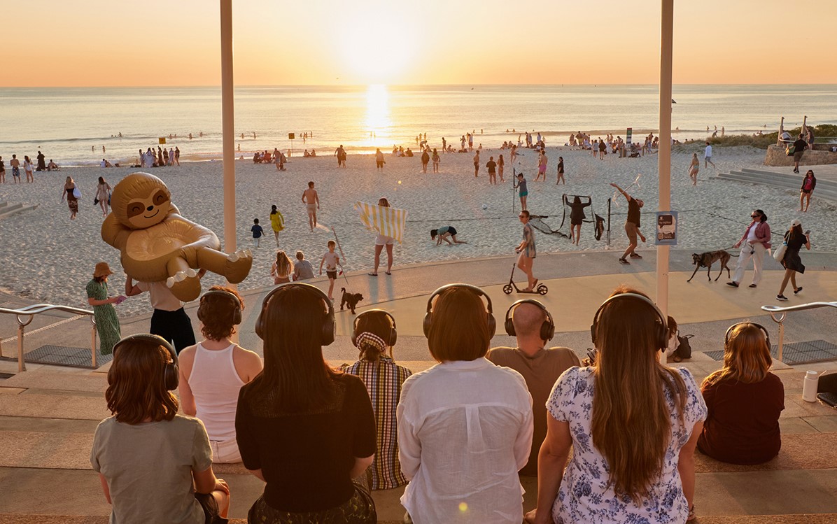 8 people sitting at an amphitheatre with headphones on looking out onto a beach with the sun setting. Beach goers are in the middle ground doing various activities 