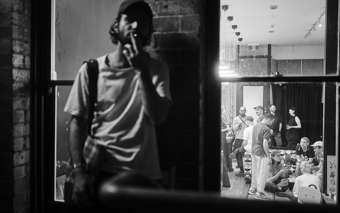 A black and white image of a performer in the foreground, slightly blurred smoking a cigarette and in focus a group of performers are talking and laughing inside. Credit Duncan Wright