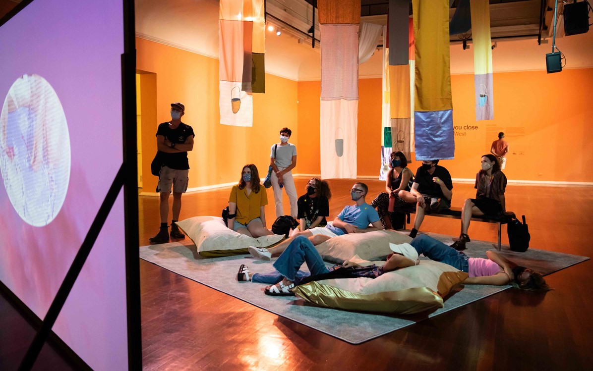 A bunch of young creatives sit on benches and beanbags watching a screen at PICA for Katie West's 'We Hold You Close' exhibition.