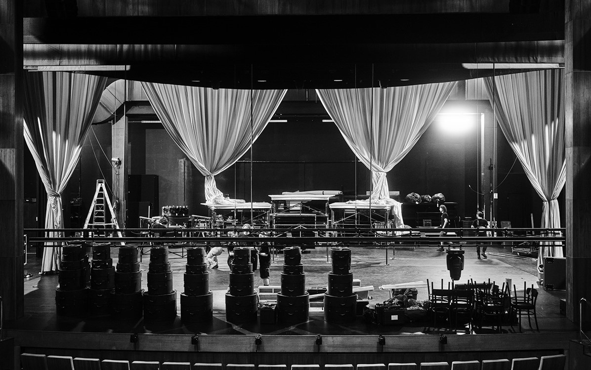 black and white image of a stage set up with boxes stacked up and chairs stacked. 4 curtains are tied up in the background.  Credit Duncan Wright