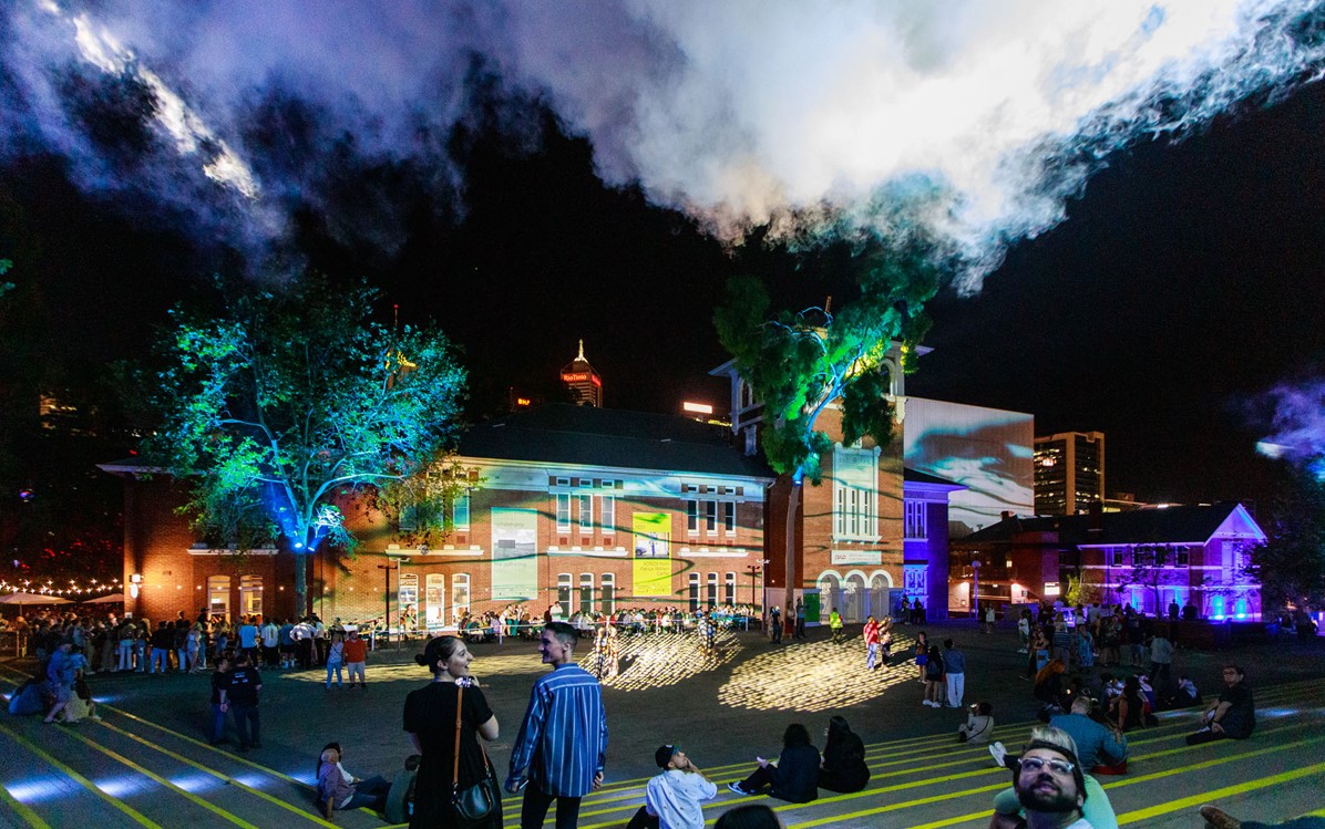 People wander through a public space, light projections appear upon a building facade and reflected in clouds and trees.