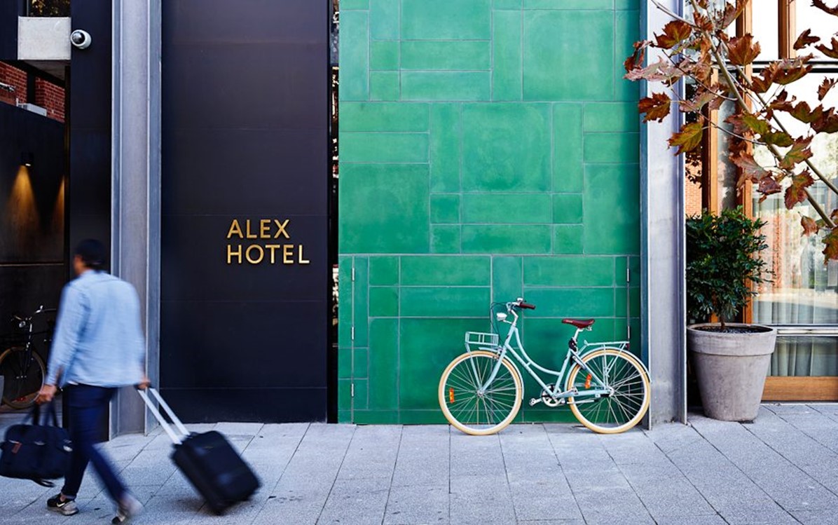 A person arrives at the Alex Hotel entry with a suitcase and bag, a bicycle leans against the green exterior wall.