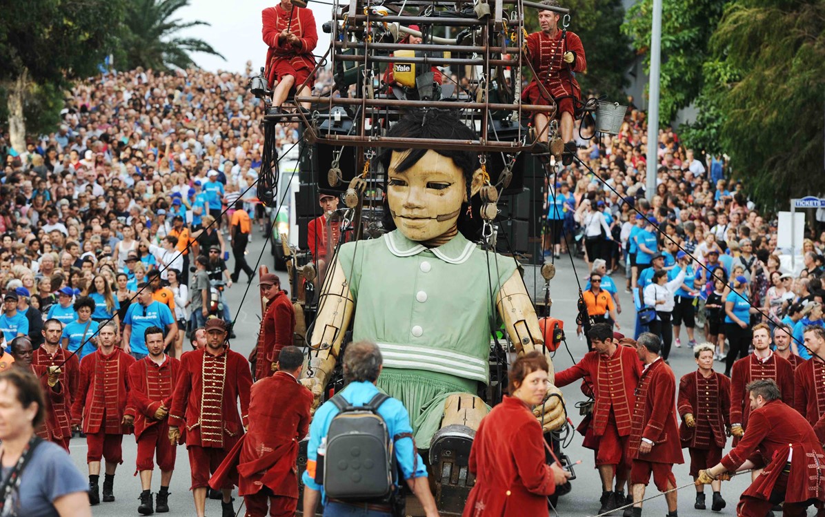 An image of a large puppet being supported and walked alongside by the performers and crowds of people are following on the street behind it