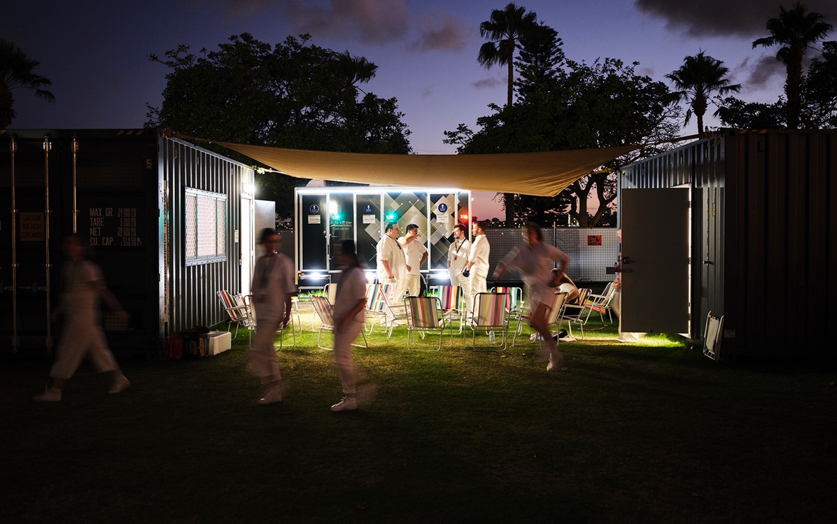 People talking in a small group, dressed in white outside at dusk. Three people are blurred in front of them, running across the grassed area in front of the others.