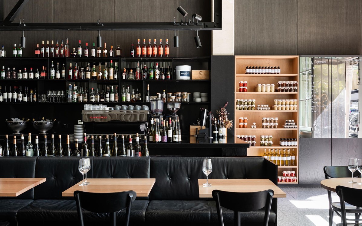 A bar/restaurant interior with black chairs and booths, and wooden tables, a fully stocked wall of bottled alcohol.