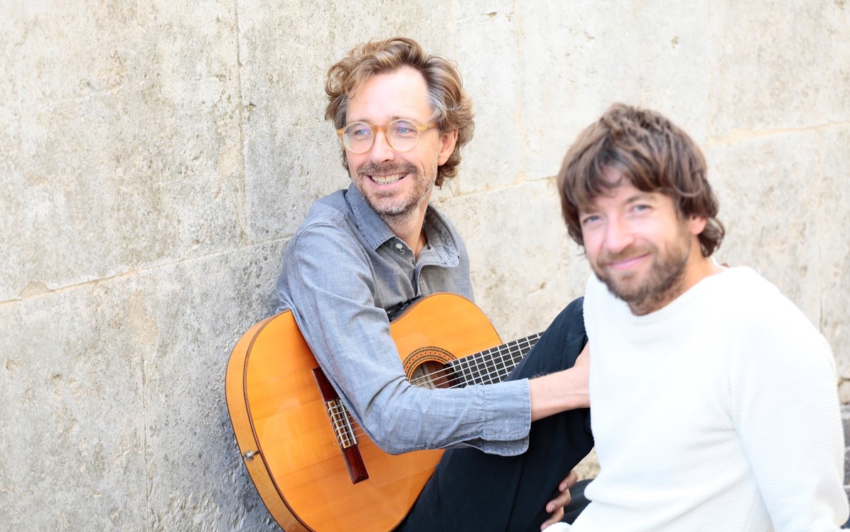 Two men sit against a concrete wall, one has glasses, both are smiling, one holds and acoustic guitar