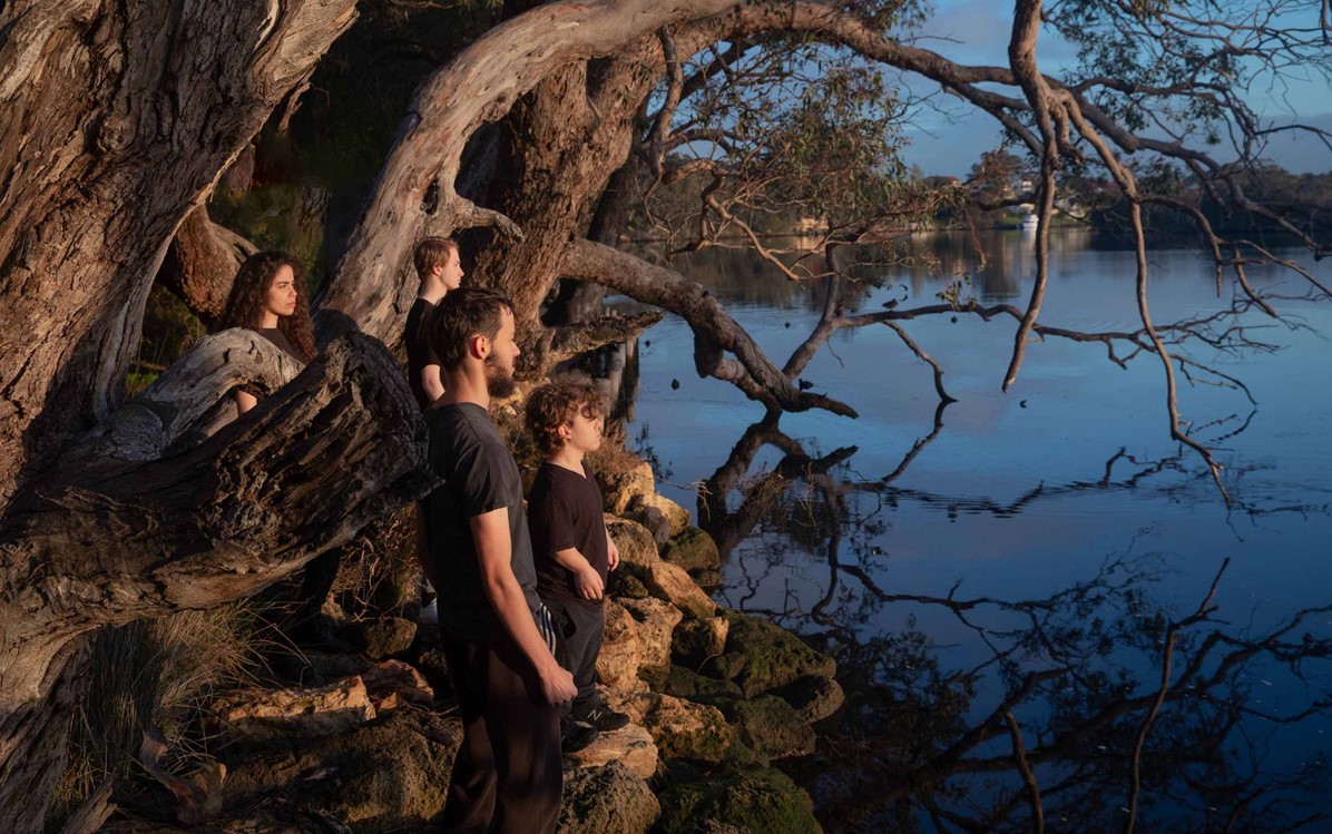  Promotional photo for theatre production "Beside". A group of people are pictured on rocks and tree roots on the bank of a river.
