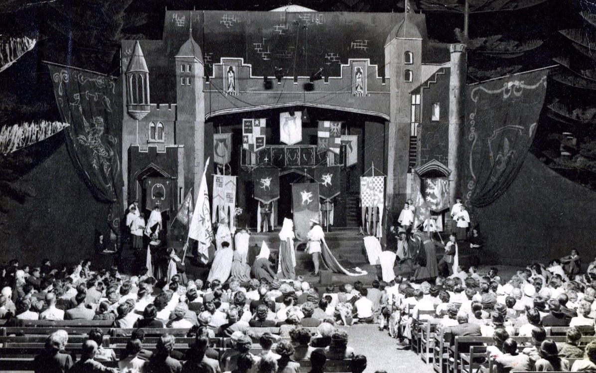 A black and white image of a medieval looking theatre performance with people seated in the audience.