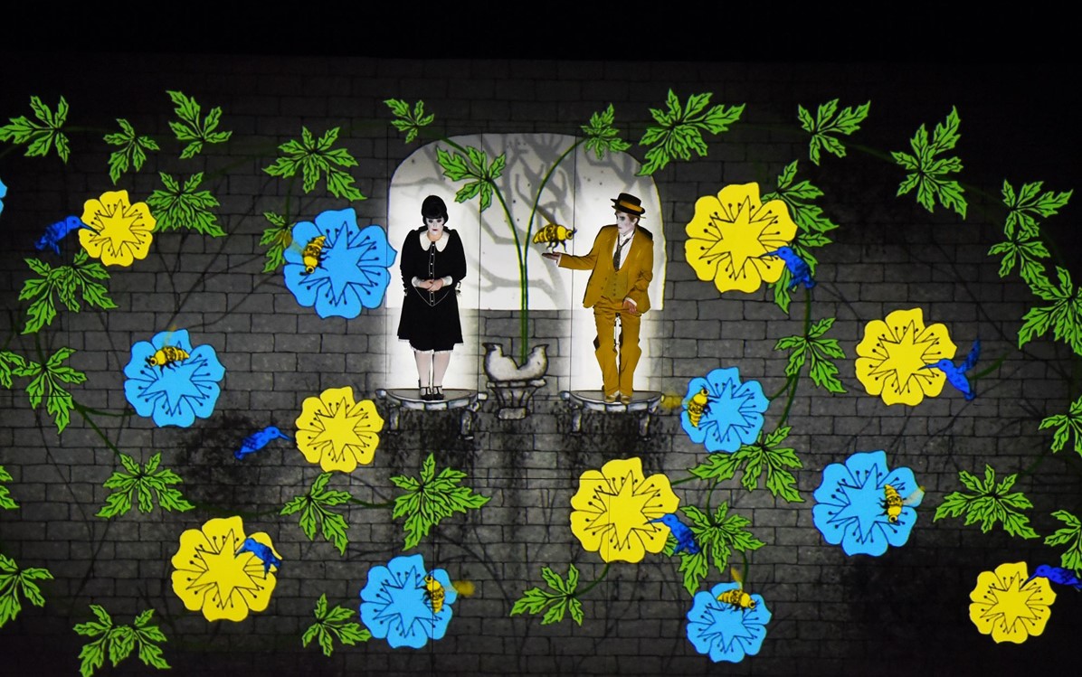 Image of the Magic Flute performance from Perth Festival 2019, two actors on stage with spotlights, blue and yellow flowers projected around them.