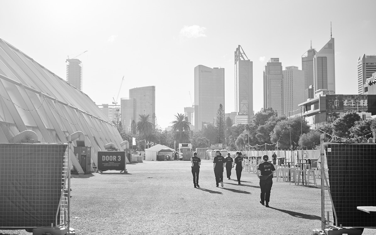 A black and white image of Perth Festival staff walking away on the grass of Langley Park. There is a big tent set up on the left hand side with a sign that says 'Door 3'.  Credit Duncan Wright