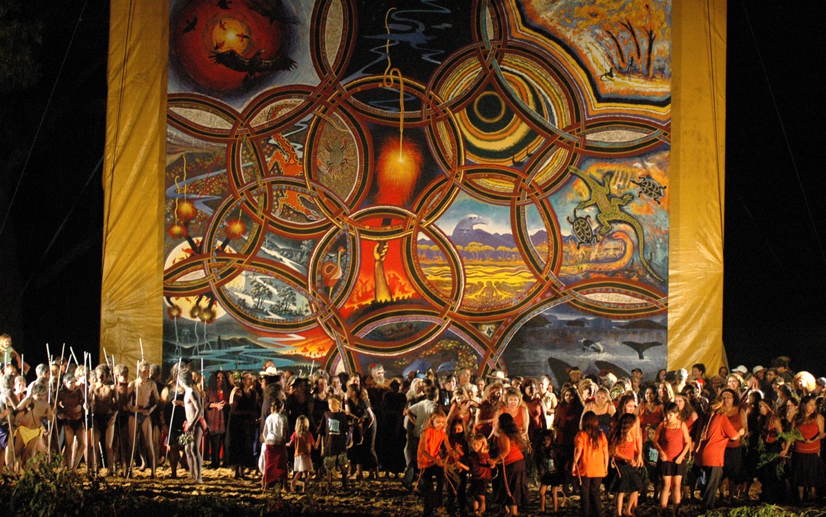 A large gathering of performers on a stage with a large artwork displayed behind them