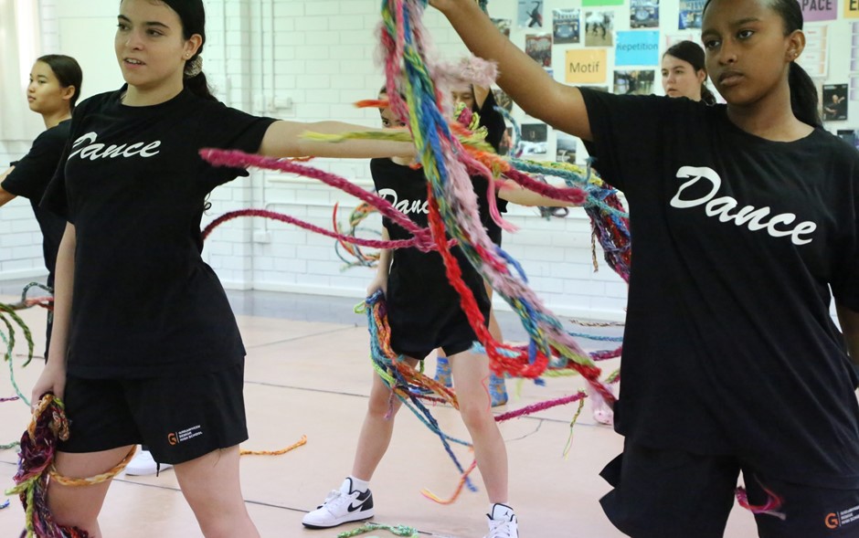 A group of young people in black shorts and black tshirts that say 'Dance' move in a room holding large bunches of knitted yarn