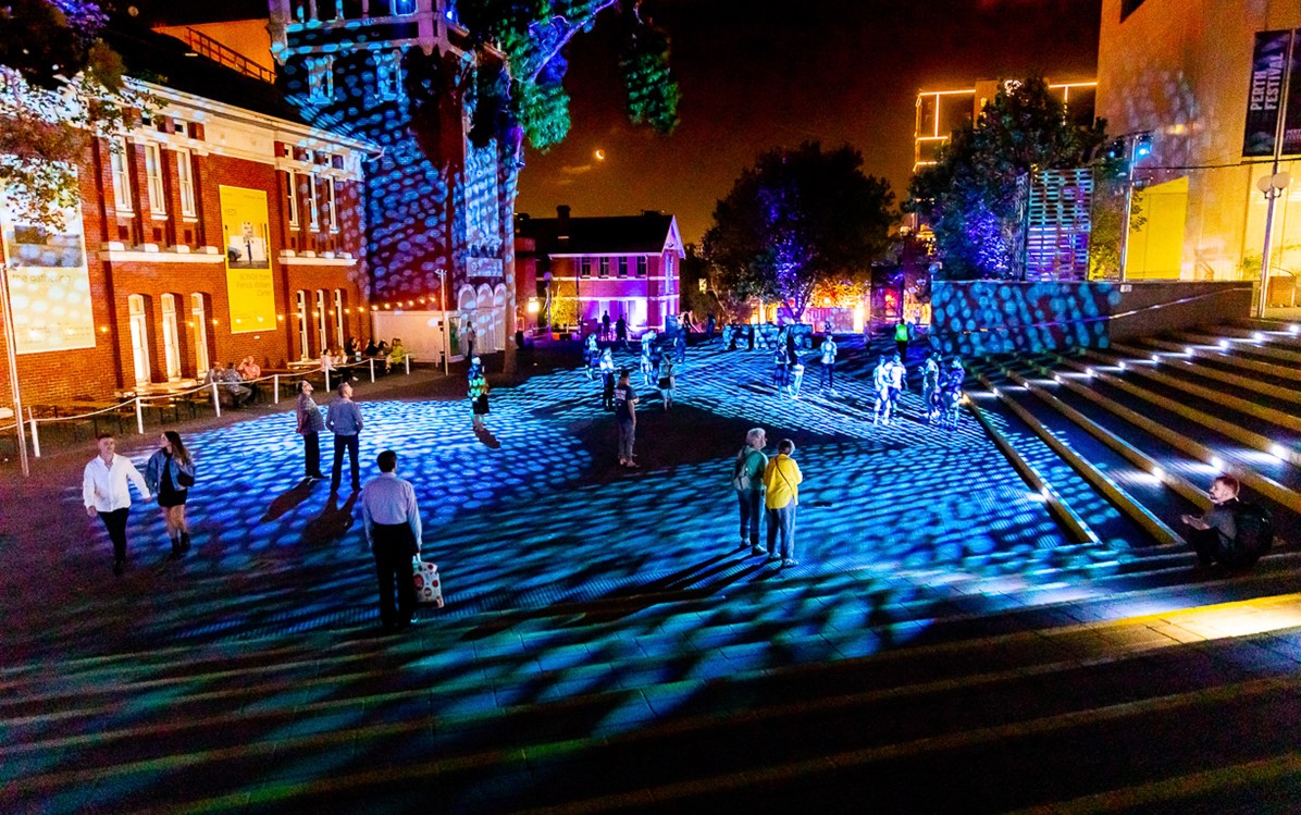 People wander around a public space at night, blue and purple light projections on the ground and surrounding buildings.