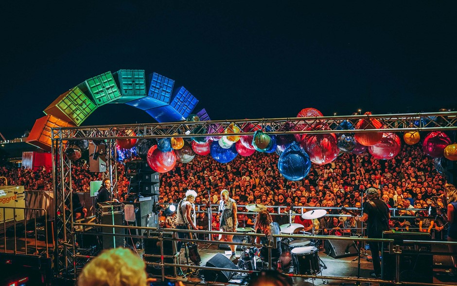 A still from Perth Festival's 2021 Highway to Hell event, a musicians play on a stage with colourful balloons, a large crowd watches on, behind is the rainbow shipping container installation in Fremantle.