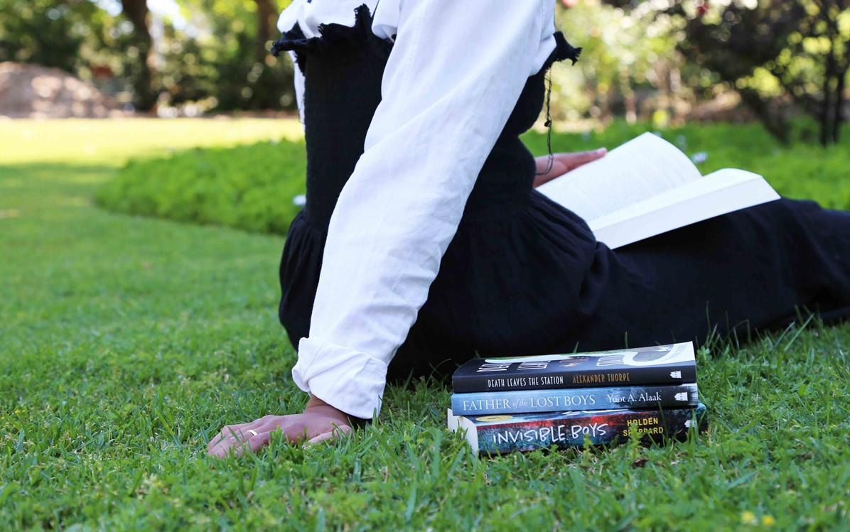 A person in a white shirt and black dress sits on grass reading a book, with a stack of three books next to them.
