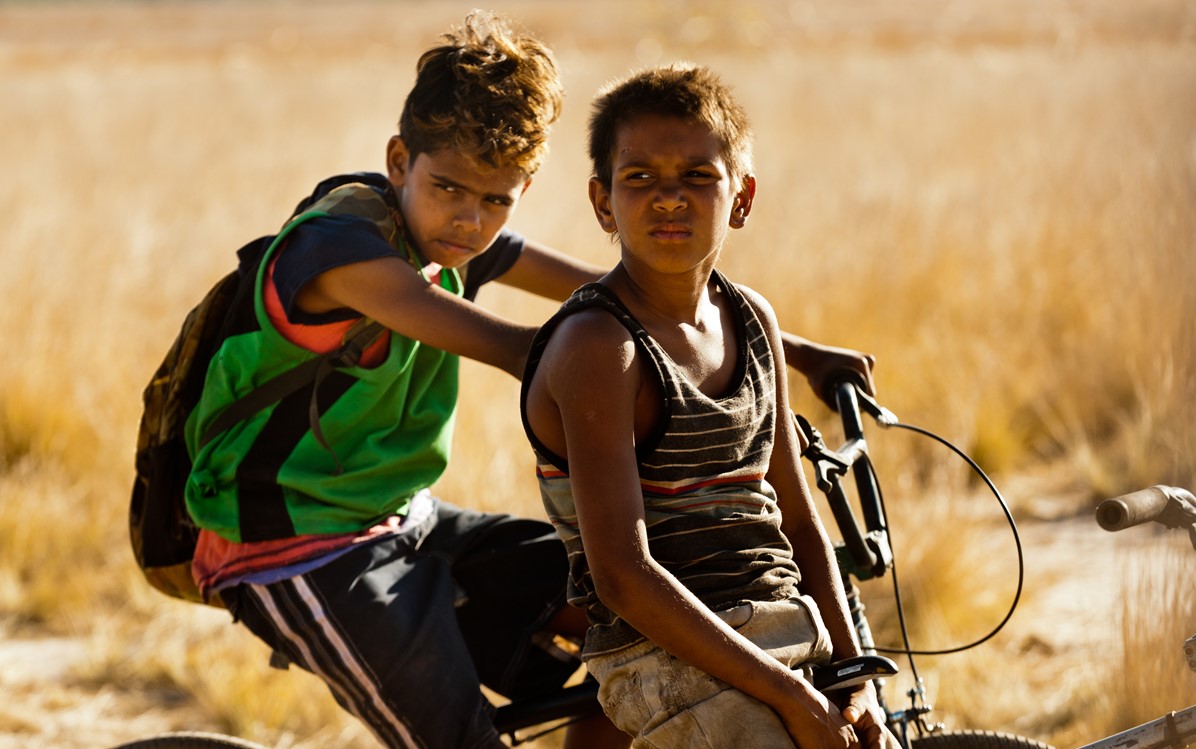 Two boys are pictured on bikes, one has a back pack. From the film "Satellite Boy".